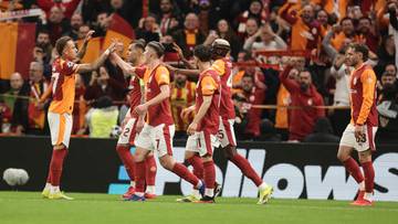 Piłkarze Galatasaray w czerwono-ż&oacute;łtych strojach świętują bramkę na boisku, z kibicami na trybunach w tle.
