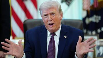 Donald Trump przemawia z otwartymi dłońmi, w garniturze i krawacie, na tle flagi USA.