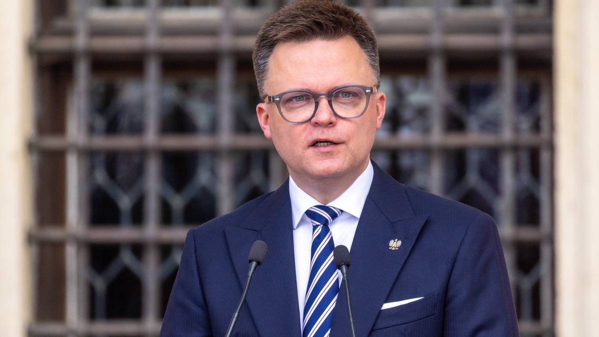 Mężczyzna w garniturze i okularach przemawia do mikrofonu.