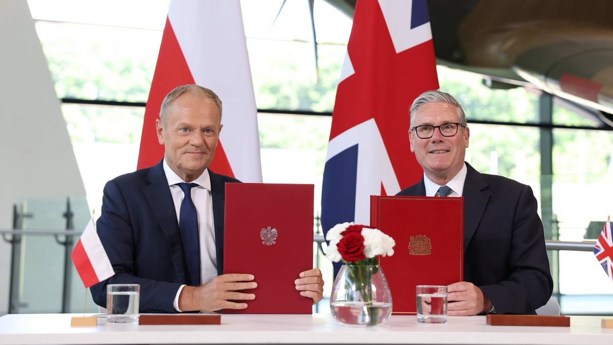 Dw&oacute;ch mężczyzn w garniturach stoi za stołem, trzymając dokumenty. Na stole stoją szklanki z wodą i bukiet kwiat&oacute;w. W tle widoczne są flagi Polski i Wielkiej Brytanii oraz fragment samolotu.