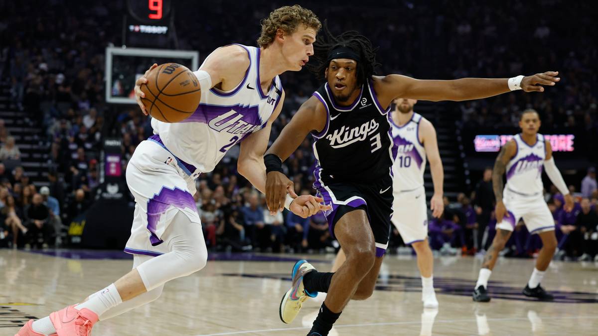 Dwóch koszykarzy podczas meczu. Zawodnik w białym stroju z fioletowymi akcentami prowadzi piłkę, a zawodnik w czarnym stroju z napisem "Kings" próbuje go zatrzymać.