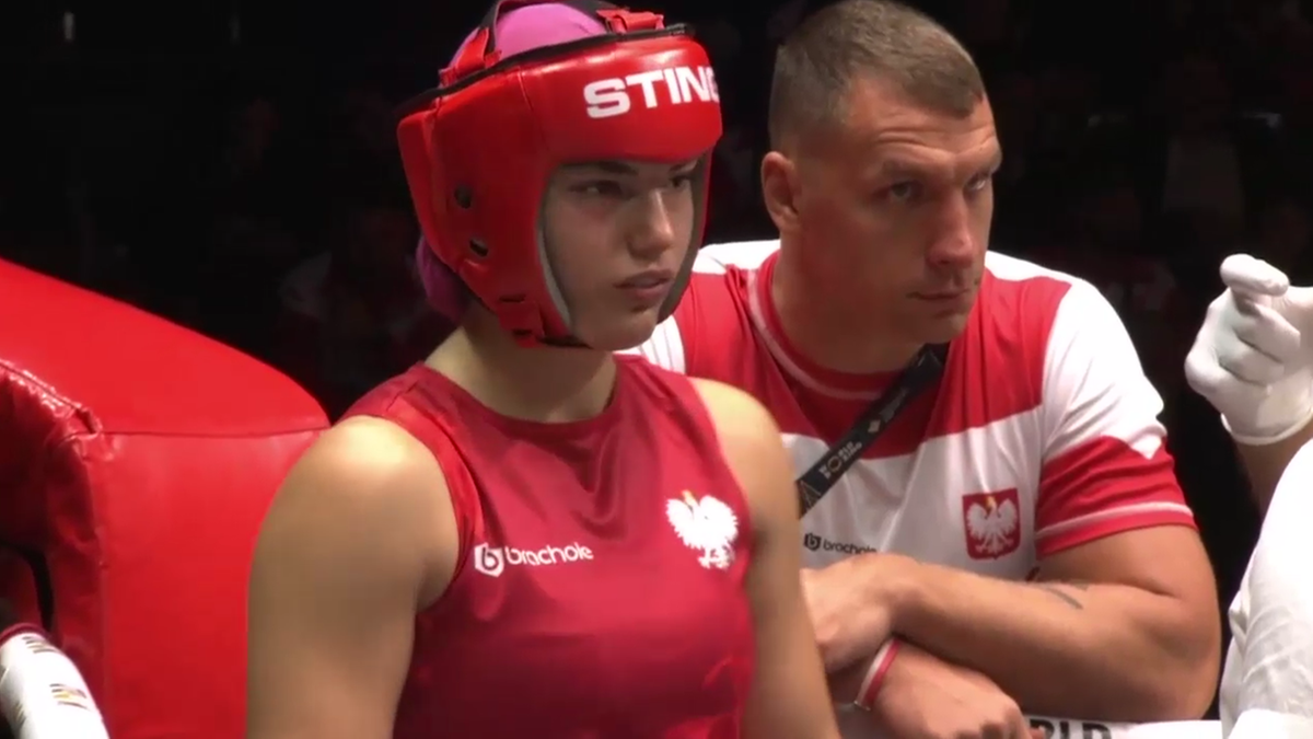 Młoda kobieta w czerwonym kasku bokserskim z białym orłem i napisem "STING" oraz w czerwonej kamizelce z białym orłem, stoi w ringu. Obok niej stoi mężczyzna w czerwono-białej koszulce z polskim orłem, który wydaje się być trenerem. Jej twarz wyraża skupienie lub smutek.