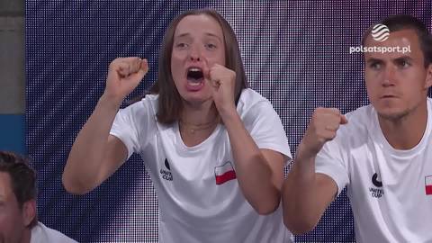 Iga Świątek z otwartymi ustami i zaciśniętymi pięściami, wyrażająca radość i ekscytację podczas meczu. Obok niej siedzi mężczyzna w polskiej koszulce.