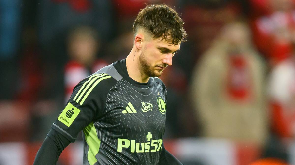 Bramkarz Legii Warszawa w zielono-szarym stroju z piłką przy nodze na boisku.