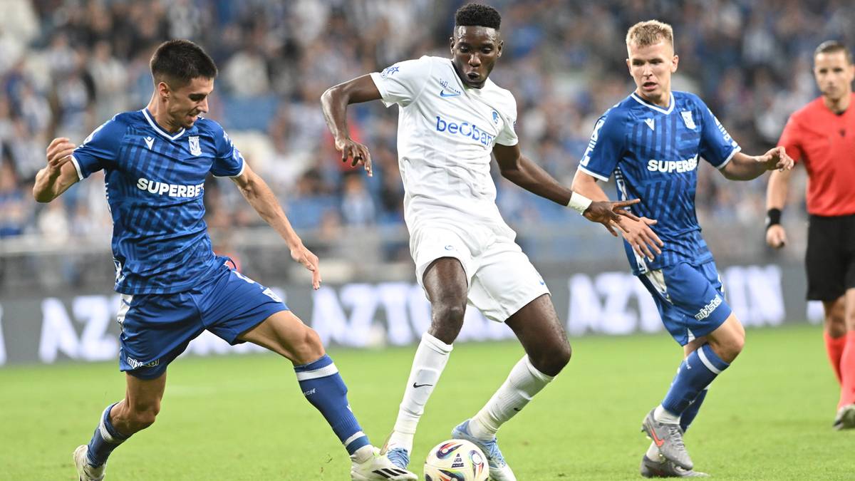 Trzech piłkarzy na zielonej murawie stadionu podczas meczu. Dwóch w niebieskich strojach próbujących odebrać piłkę graczowi w białym stroju.