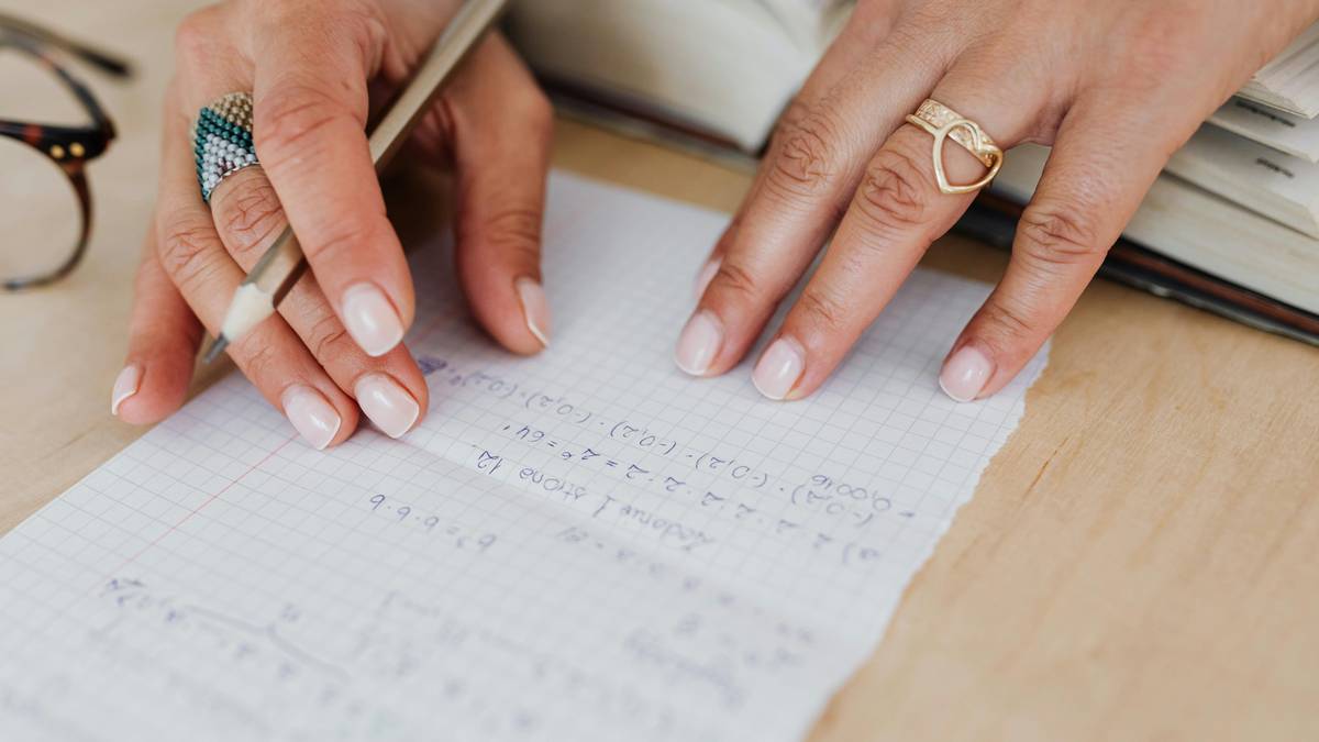 Dłonie osoby z pierścionkami piszącej ołówkiem na kartce w kratkę.