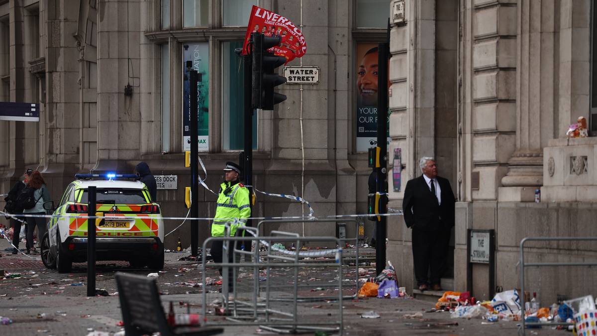 Funkcjonariusz policji w jaskrawożółtej kamizelce stoi obok policyjnego radiowozu z włączonymi światłami. Wokół widoczne jest policyjne taśmy, śmieci i zniszczenia.