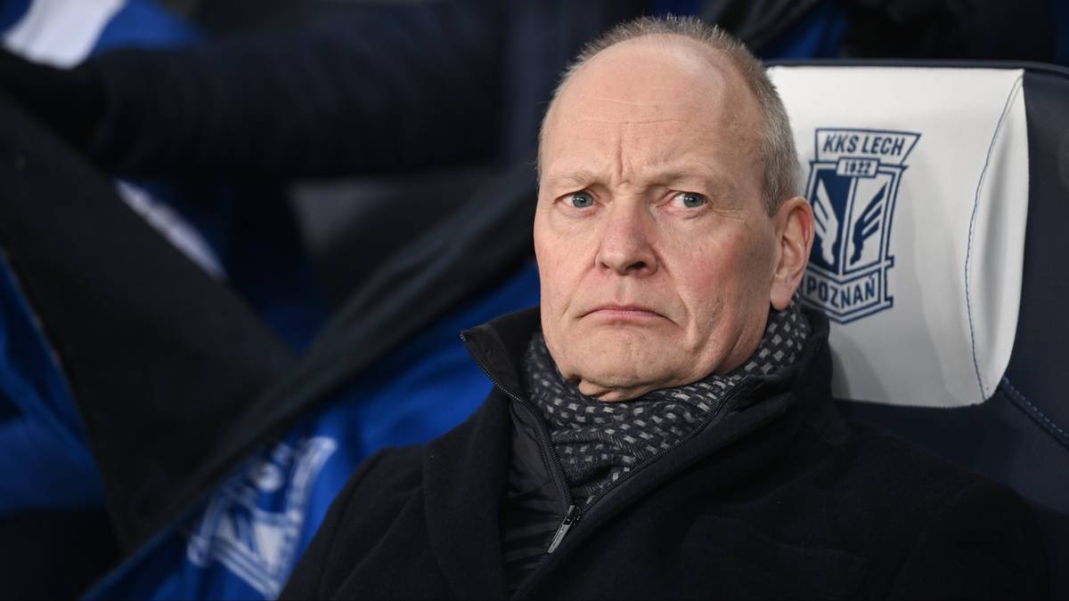 Starszy mężczyzna siedzi na ławce rezerwowych, ubrany w ciemny płaszcz i szalik, z poważnym wyrazem twarzy.