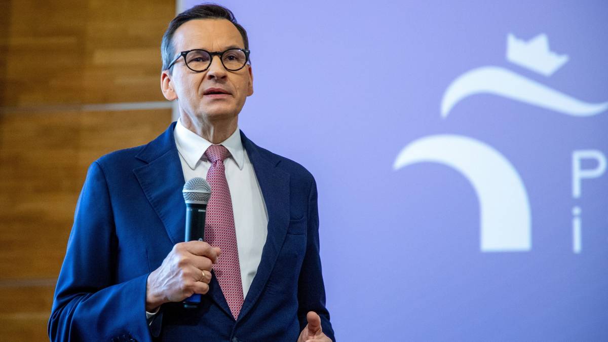 Mateusz Morawiecki przemawia do mikrofonu na tle niebieskiej ściany z logo.