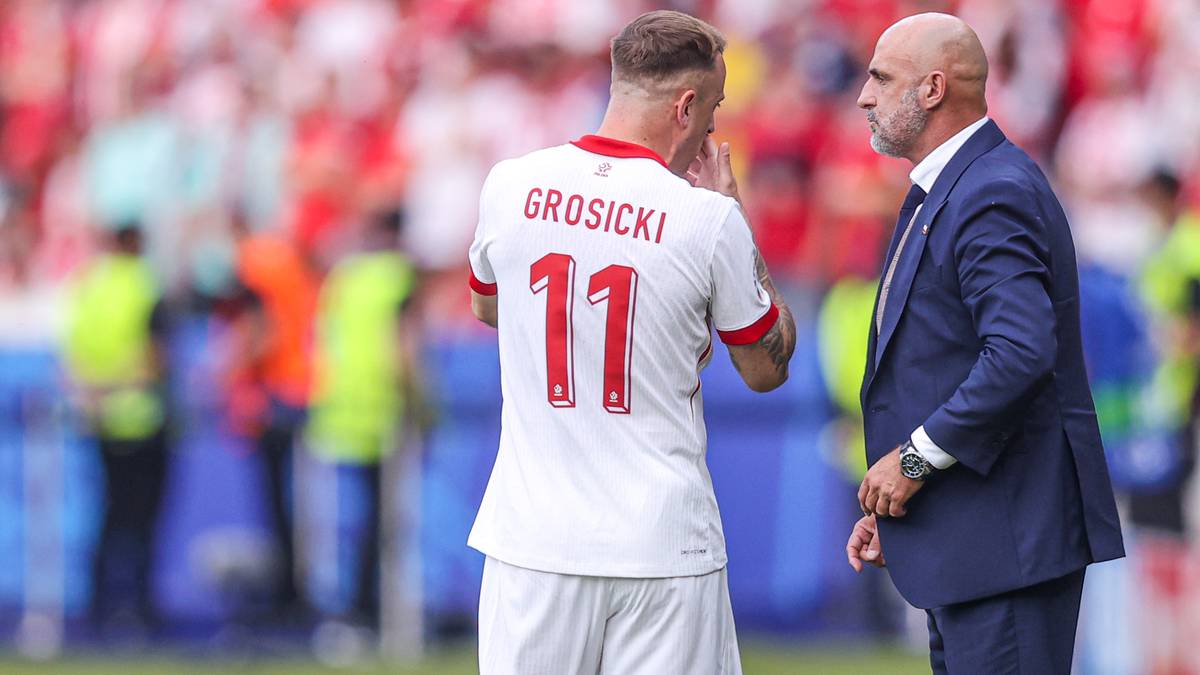 Piłkarz Kamil Grosicki z numerem 11 na plecach w białej koszulce i trener z siwą brodą w granatowym garniturze rozmawiają na boisku.