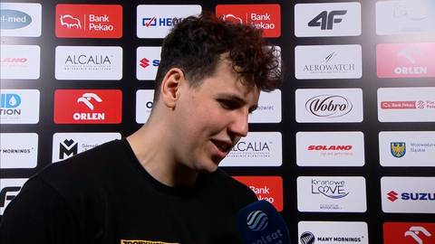 Młody mężczyzna z kręconymi włosami stoi przed ścianą z logo sponsor&oacute;w i uśmiecha się.