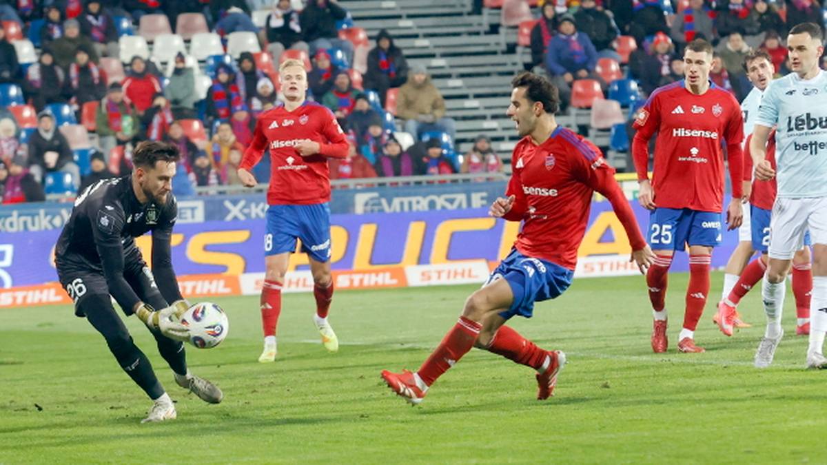 Piłkarz w czarnym stroju próbuje odebrać piłkę piłkarzowi w czerwonym stroju na zielonej murawie stadionu.