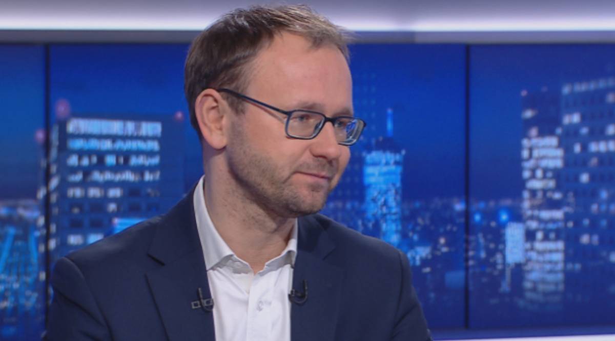 Mężczyzna w okularach i ciemnym garniturze siedzi i m&oacute;wi.