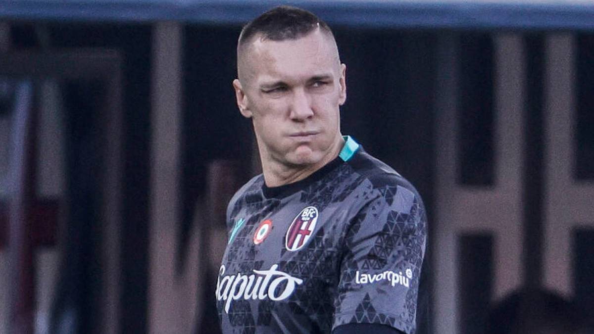 Bramkarz Łukasz Skorupski w czarnej koszulce i rękawicach bramkarskich stoi na boisku piłkarskim, obok niego idzie mężczyzna w niebieskiej kurtce.