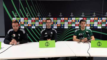 Trzech mężczyzn siedzi za stołem podczas konferencji prasowej. Dw&oacute;ch z nich to piłkarze Legii Warszawa, ubrani w stroje klubowe. Trzeci mężczyzna to trener, r&oacute;wnież w stroju klubowym. Wszyscy siedzą przed mikrofonami, a za nimi znajduje się ściana z logo UEFA Europa League oraz sponsor&oacute;w.