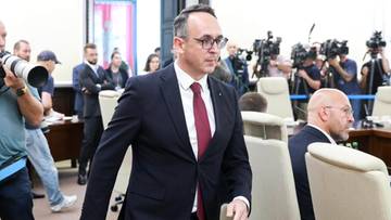 Polityk w garniturze idący przez salę w otoczeniu dziennikarzy i kamer.
