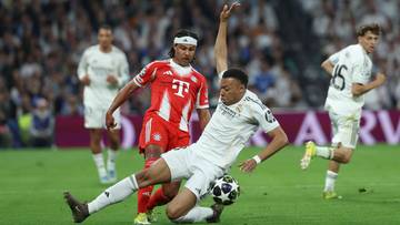 Piłkarze w białych strojach rywalizują o piłkę na zielonej murawie stadionu, w tle widać innych zawodnik&oacute;w i trybuny.