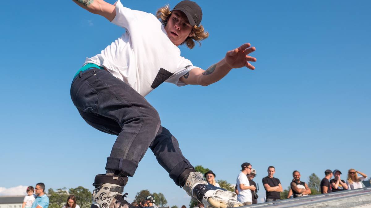 Młody mężczyzna w białej koszulce i czapce z daszkiem wykonuje ewolucję na rolkach, jego ciało jest w ruchu, a ręce są rozłożone na boki. W tle widać grupę obserwatorów.