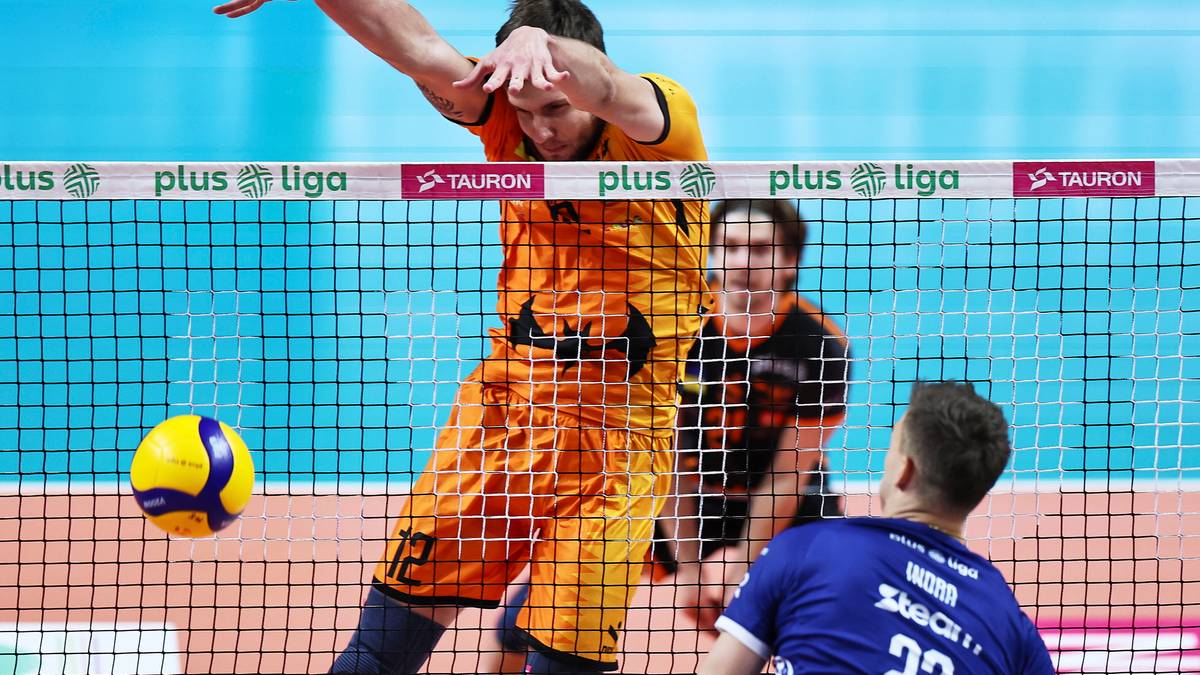 Siatkarz w pomarańczowym stroju wykonuje wysoki skok do ataku, z rękami wyciągniętymi w kierunku siatki. Obok niego widać piłkę do siatkówki. Po drugiej stronie siatki stoją dwaj zawodnicy w czerwonych i niebieskich strojach.