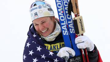 Uśmiechnięta narciarka biegowa Jessica Diggins owinięta flagą USA, trzymająca narty i kijki.