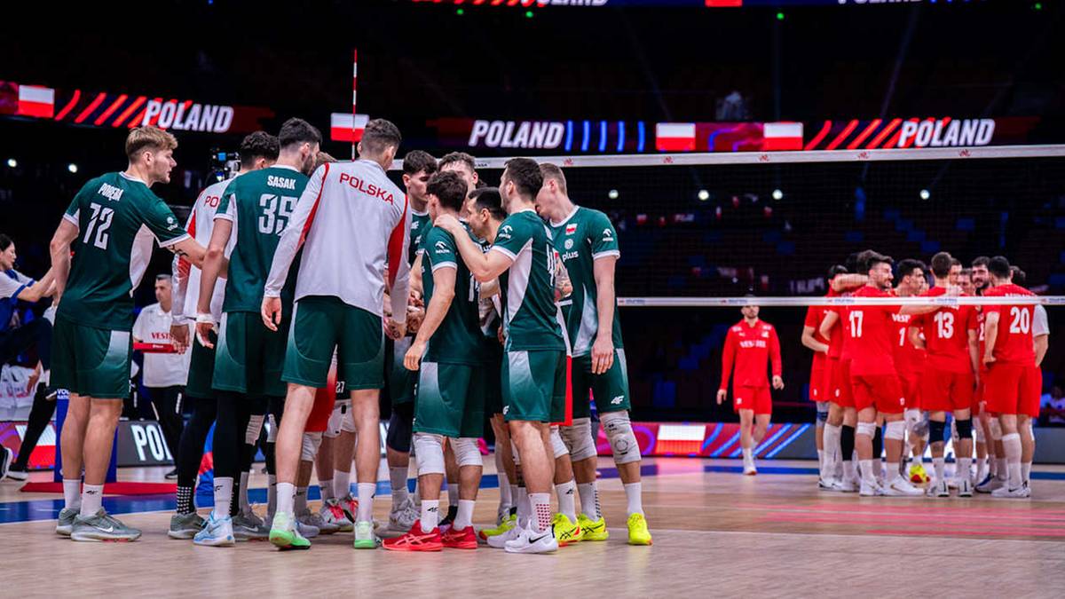 Drużyna siatkarzy w zielonych strojach stoi blisko siebie, skupiona i w środku dyskusji z trenerem. W tle widać drugą drużynę w czerwonych strojach.