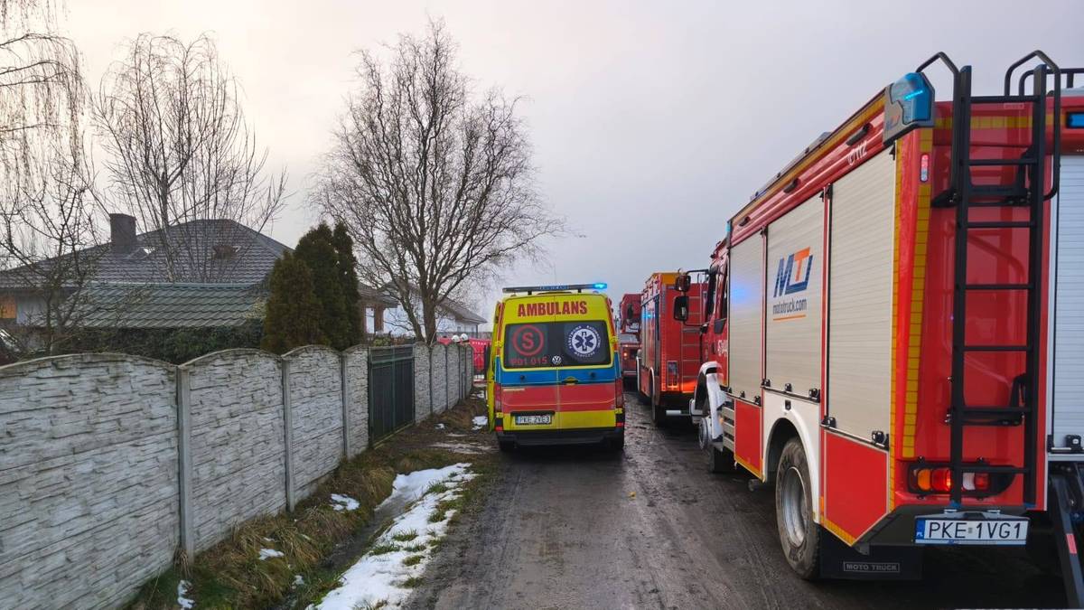 Samochody strażackie i karetka pogotowia stoją na drodze w pobliżu dom&oacute;w.