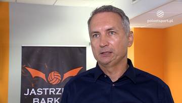 Trener Andrzej Kowal wypowiadający się na tle ciemnego rollupa z logo klubu i pomarańczowej ściany.
