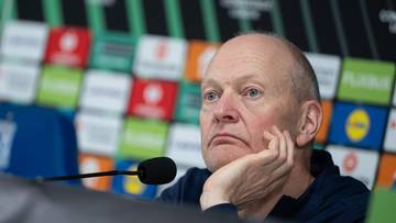 Mężczyzna w średnim wieku z łysiną, oparty podbródkiem o dłoń, patrzy w bok z lekko zamyślonym wyrazem twarzy. W tle widoczna ściana z kolorowymi logotypami.