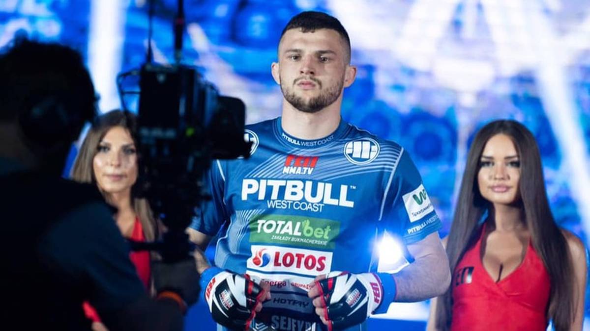 Cezary Oleksiejczuk, zawodnik MMA, w stroju sportowym z rękawicami bokserskimi, stoi na tle rozmazanych postaci i świateł.