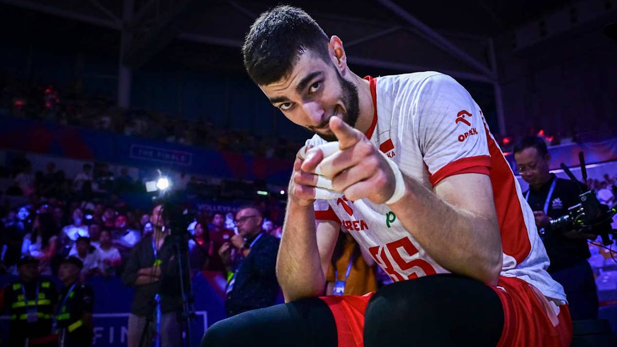 Siatkarz w białej koszulce z czerwonymi wstawkami wskazuje palcem na widza, uśmiechając się.