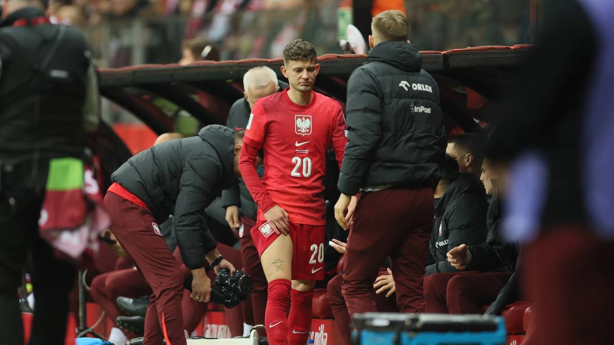 Piłkarz reprezentacji Polski w czerwonej koszulce z numerem 20, stojący na boisku, opuszcza murawę, otoczony przez innych zawodników i członków sztabu.