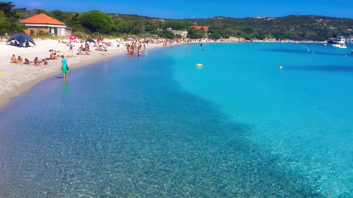 Plaża z turkusową wodą i piaszczystym brzegiem, na kt&oacute;rym odpoczywa wielu ludzi.