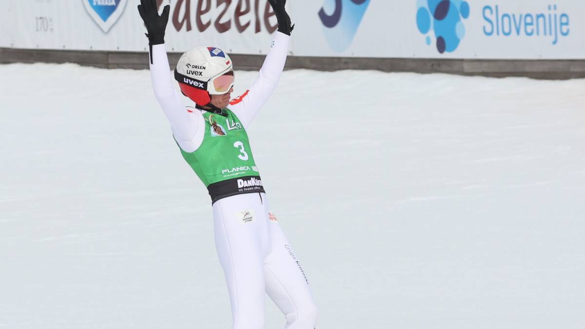 Kamil Stoch świętuje udany skok narciarski, z rękami uniesionymi w g&oacute;rę, na tle trybun z kibicami.