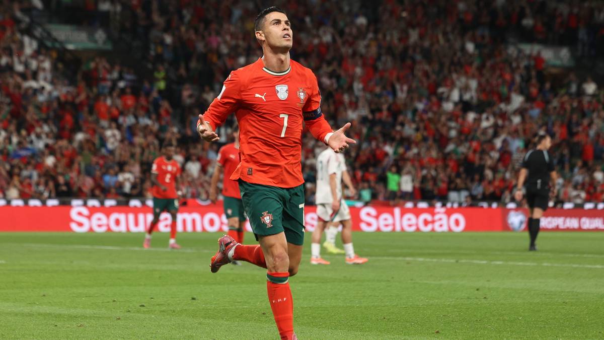 Cristiano Ronaldo w pomarańczowej koszulce piłkarskiej z numerem 7, radośnie celebruje bramkę na boisku, z trybunami pełnymi kibiców w tle.