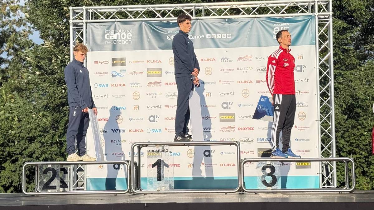 Trzech młodych mężczyzn stoi na podium sportowym. Pierwszy z lewej, w niebieskim dresie, na trzecim stopniu podium. Drugi, w granatowym dresie, na najwyższym stopniu podium. Trzeci, w czerwonej kurtce, na drugim stopniu podium.