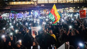 Tłum ludzi trzyma w g&oacute;rze światła telefon&oacute;w podczas nocnej demonstracji, na kt&oacute;rej widoczna jest tęczowa flaga.