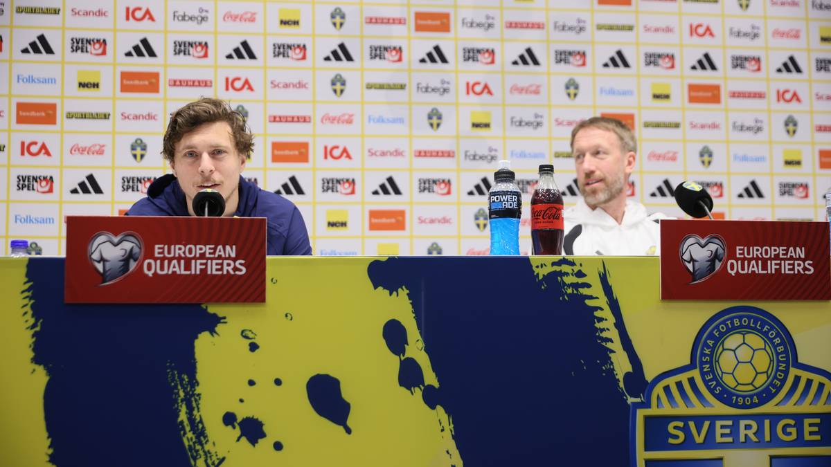 Dwóch mężczyzn siedzi przy stole konferencyjnym przed ścianą z logotypami. Jeden z nich, młodszy, mówi do mikrofonu, drugi, starszy, patrzy w bok.