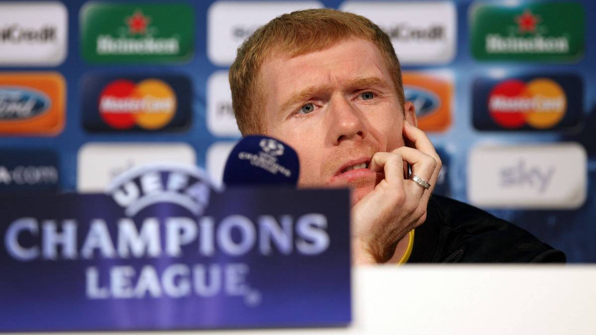 Mężczyzna o rudych włosach i jasnej karnacji, w czarnej koszulce, siedzi za mikrofonem i tablicą z logo Ligi Mistrzów UEFA. Jest w trakcie konferencji prasowej, trzymając palce przy uchu.