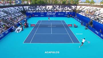 Kort tenisowy podczas meczu z widokiem z góry. Na niebieskiej nawierzchni rozgrywa się gra między dwoma zawodnikami. Wokół kortu znajdują się trybuny z widzami. U dołu kadru widnieje napis "ACAPULCO".