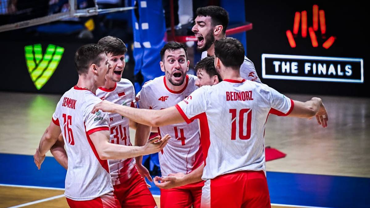 Grupa siatkarzy w białych koszulkach z czerwonymi detalami świętuje zwycięstwo na boisku. Ich twarze wyrażają radość i wzruszenie.