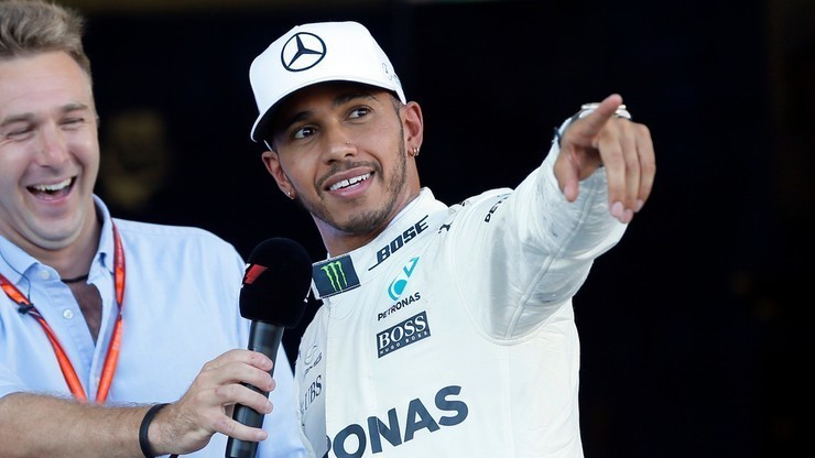 Lewis Hamilton w kombinezonie Mercedes-AMG Petronas Formula One Team, ubrany w białą czapkę z logo Mercedesa, uśmiecha się i wskazuje palcem na coś po swojej prawej stronie. Obok niego stoi mężczyzna z mikrofonem.