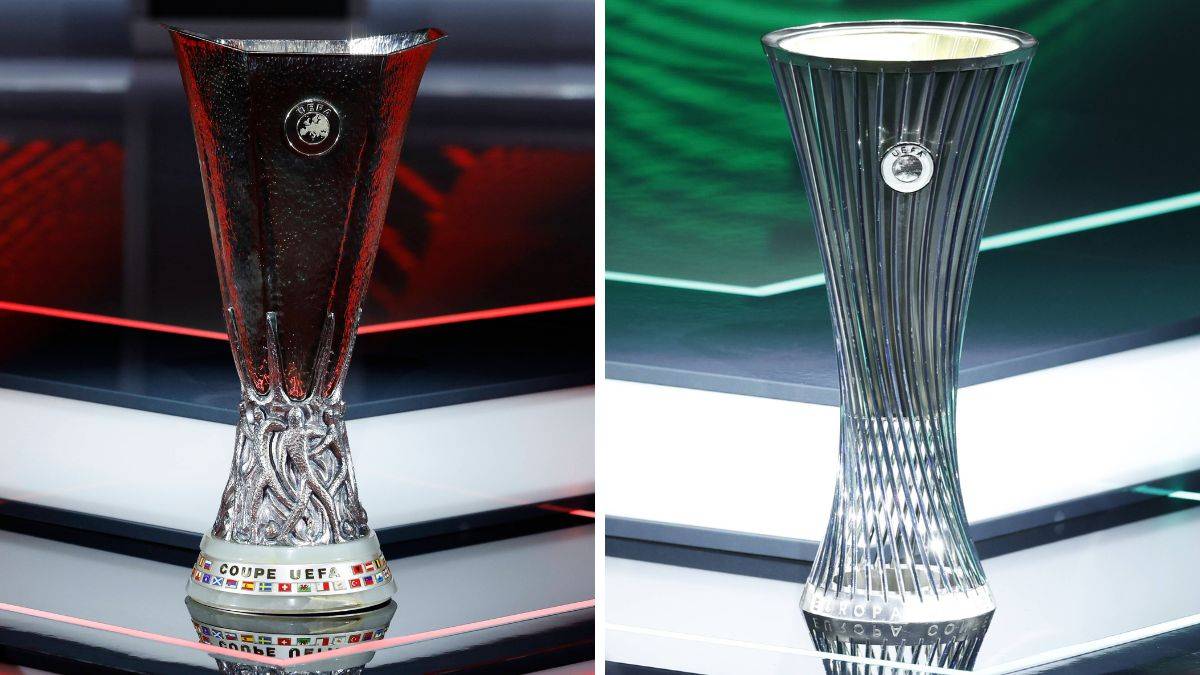 Dwa puchary UEFA ustawione obok siebie na tle stoiska z niebieskimi i zielonymi światłami. Po lewej stronie jest srebrny puchar Ligi Europy, a po prawej srebrny puchar Ligi Konferencji.
