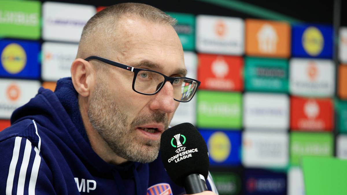 Portret mężczyzny w okularach z krótką bródką, mówiącego do mikrofonu z logo Ligi Konferencji Europy. Na jego ubraniu widoczny napis "VP". W tle kolorowe panele z logotypami.