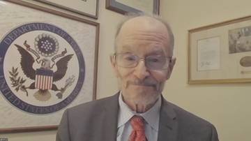 Starszy mężczyzna w okularach i garniturze z krawatem, stojący przed pieczęcią Departamentu Stanu USA.