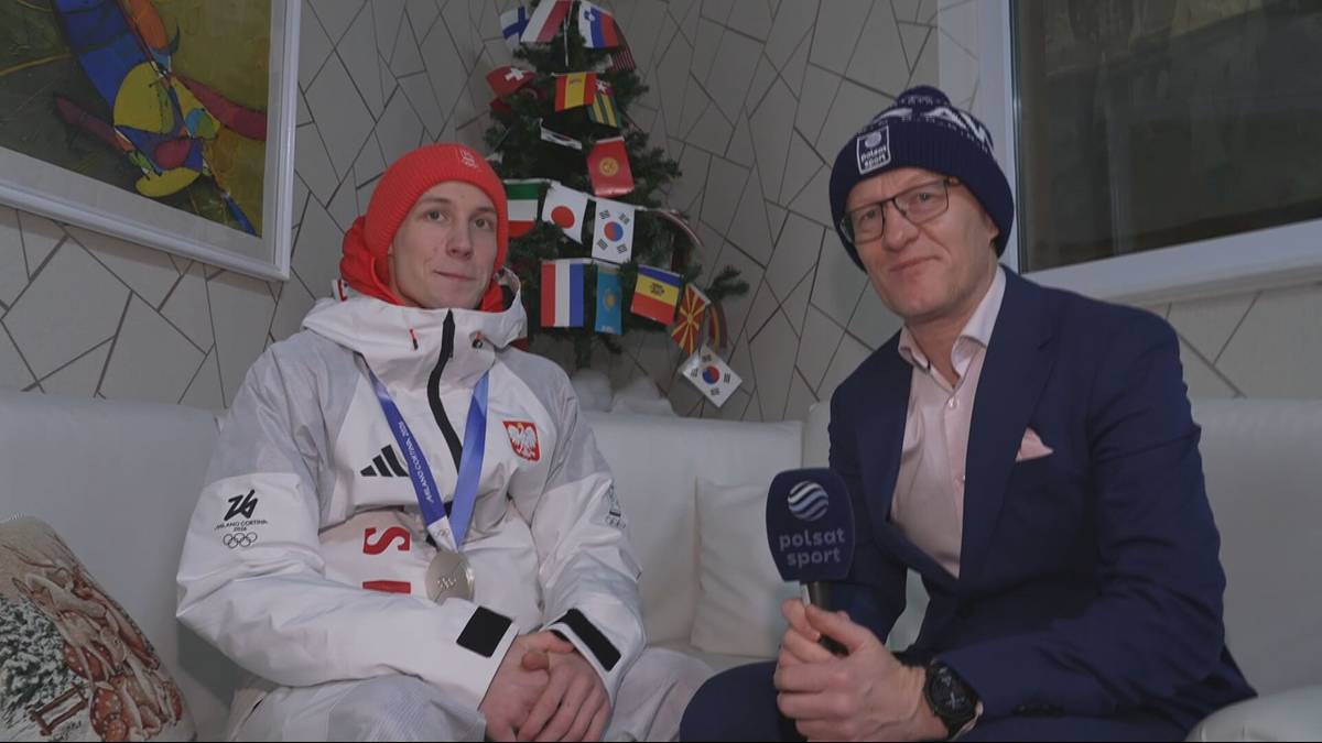 Dwóch mężczyzn siedzi na kanapie. Jeden z nich jest sportowcem w stroju zimowym z medalem, drugi to dziennikarz z mikrofonem. W tle znajduje się ozdobiona flagami choinka.