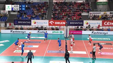 Ujęcie z meczu siatkówki, gdzie zawodnicy w niebieskich i czerwonych strojach rywalizują pod siatką. Na ekranie widoczny jest wynik 14:15.
