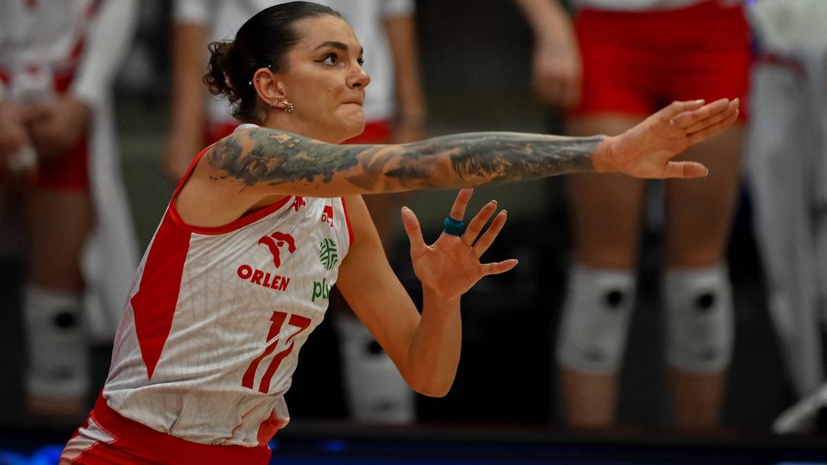 Siatkarka w białej koszulce z czerwonymi wstawkami, numerem 17 i logo, wykonująca akcję z piłką. Ma krótkie ciemne włosy związane w kucyk, widoczne tatuaże na ramieniu.