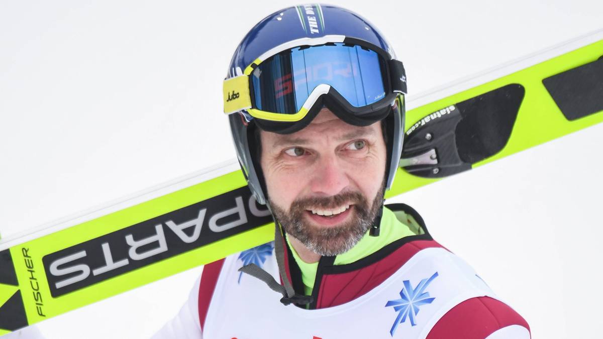 Zbliżenie na twarz mężczyzny w kasku narciarskim i goglach narciarskich, z nartami narciarskimi opartymi na ramieniu.