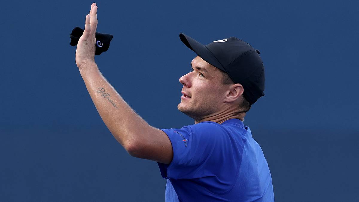 Tenisista Krystian Majchrzak w niebieskiej koszulce i czapce z daszkiem pozdrawia publiczność, unosząc prawą rękę.