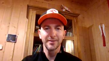 Mężczyzna w czapce z daszkiem i bluzie patrzy w obiektyw kamery. Na ścianie za nim widać drewniane panele, drzwi szafy, tablet i dekoracje.